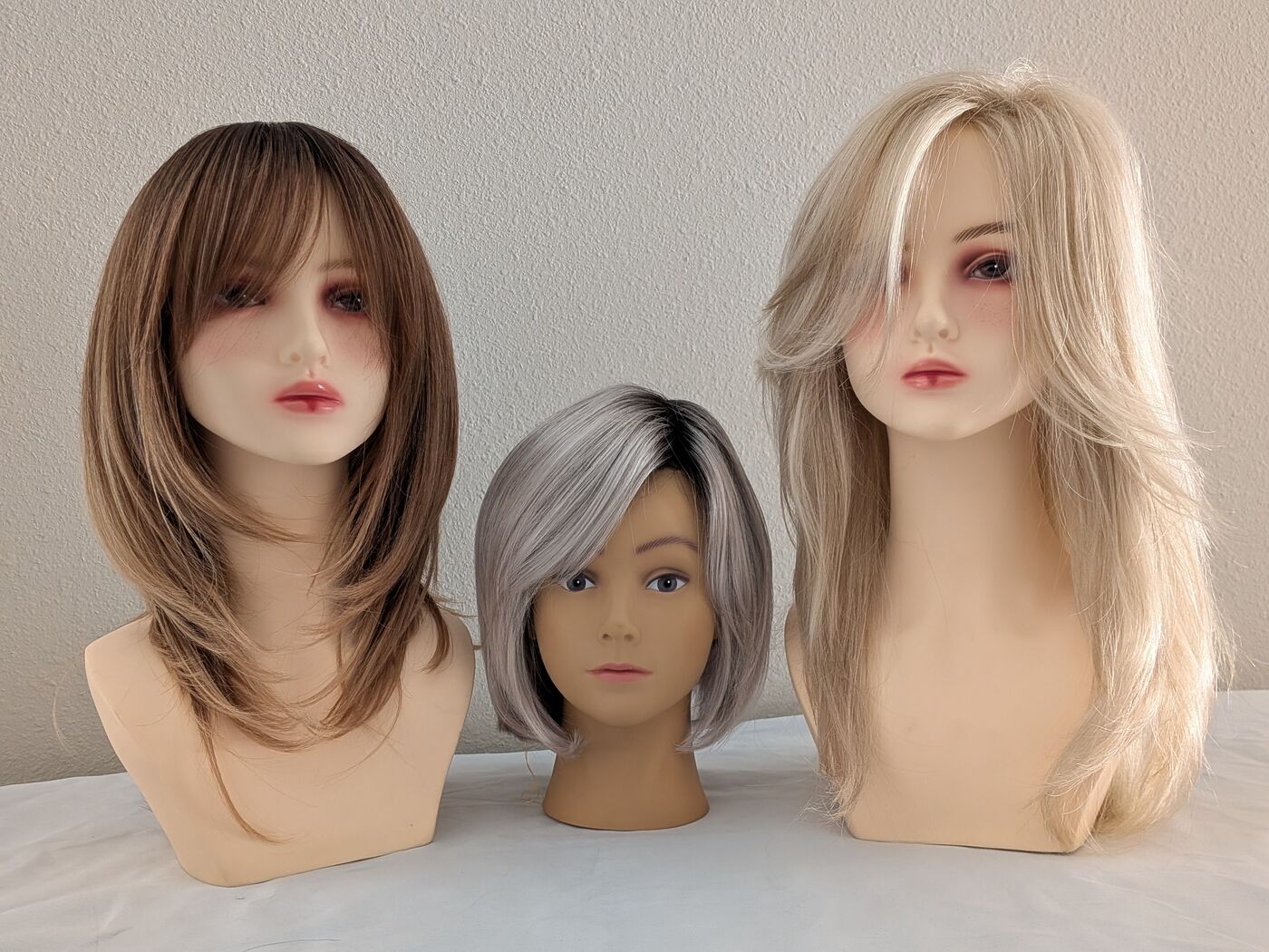 Three wigs in warm brunette, silver, and blonde styled on mannequin heads.